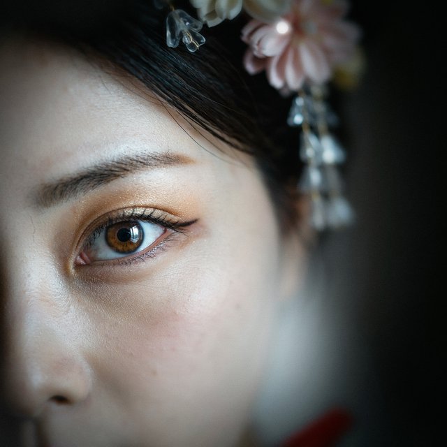 Extreme close-up macro photography of a Geisha's face. The focus is on her eyes, revealing intense emotion and hyper-realistic detail. Her skin is covered in traditional white 'Oshiroi' makeup, showing subtle, realistic cracking and skin texture. Dramatic Chiaroscuro lighting with deep shadows and a single soft light source highlighting the shimmer in her golden eyeshadow and the wetness of her eyes. Ornate kanzashi (hair ornaments) visible in the blurred periphery with intricate silk flowers and dangling silver petals. Cinematic composition, shot on 100mm macro lens, incredibly detailed eyelashes and iris patterns. — Model 7