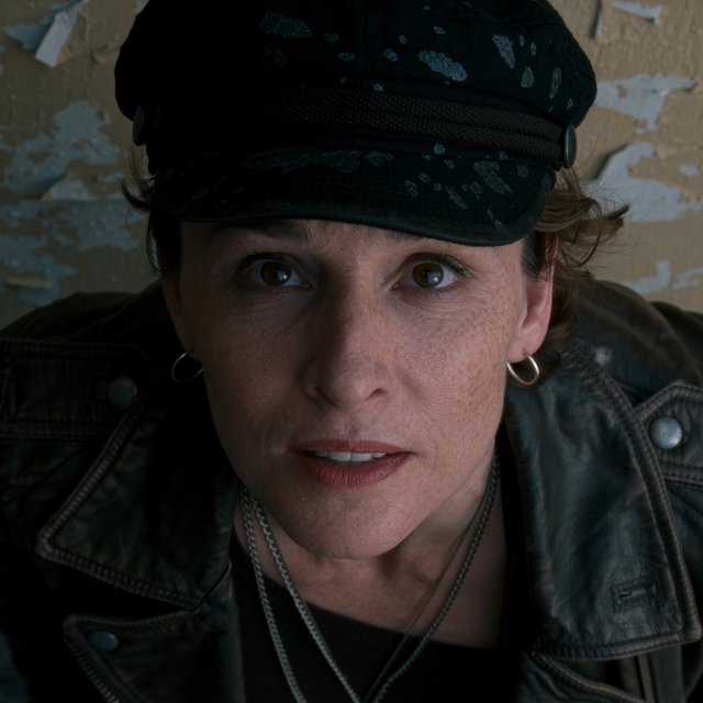 A monochrome, grainy, photorealistic medium close-up shot from a high angle: a young woman with messy, short curls, fair, freckled skin, and an intense, direct gaze into the camera. She is wearing a faded, worn-in black flat cap, a distressed vintage leather jacket over a dark undershirt, several thin silver chains, and a small hoop earring. Soft, diffused side lighting from the left casts gentle shadows across her face. In the background, a weathered wall with peeling plaster. Shallow depth of field, a moody, gritty film aesthetic, and sharp focus on the subject's eyes. — Kling v3 Omni