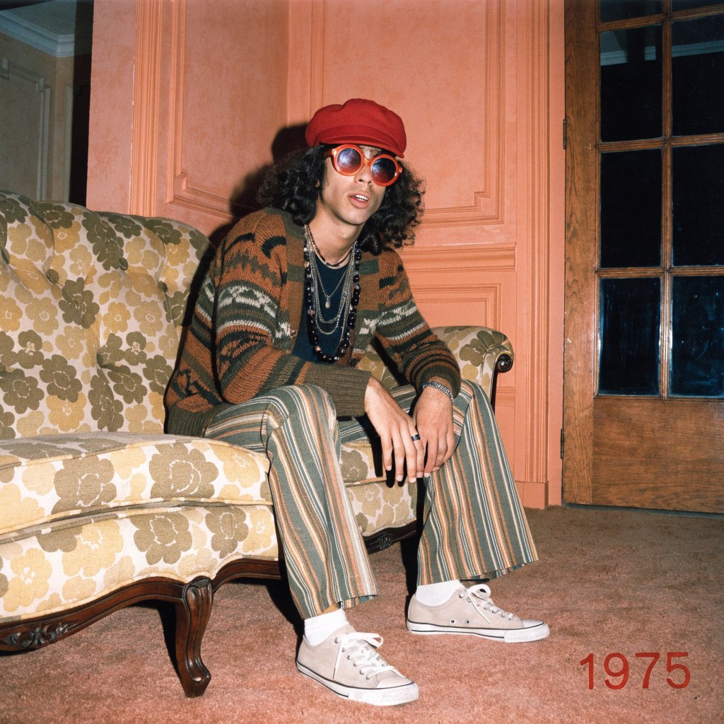 Create an image of a man in the quintessential style of 1975. Style his hair, clothing, accessories, and surroundings to reflect the distinctive look of the mid-1970s—featuring bold silhouettes, eye-catching accessories, layered details, and characteristic colors and textures. Give the image the feel of an authentic 1975 photograph, complete with analog film grain, faded colors, direct flash lighting, and a slight soft-focus effect. Add a period-accurate, red-orange date stamp in a 1970s style in one of the bottom corners. No recognizable posters. No modern objects or text. — Recraft v4