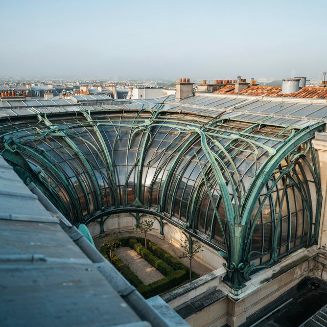 From a nearby rooftop, an Art Nouveau conservatory curves around a planted courtyard, its green copper ribs, clear glass, and pale stone base catching hazy morning light; orderly rooflines and believable supports show from above. — Ideogram v4