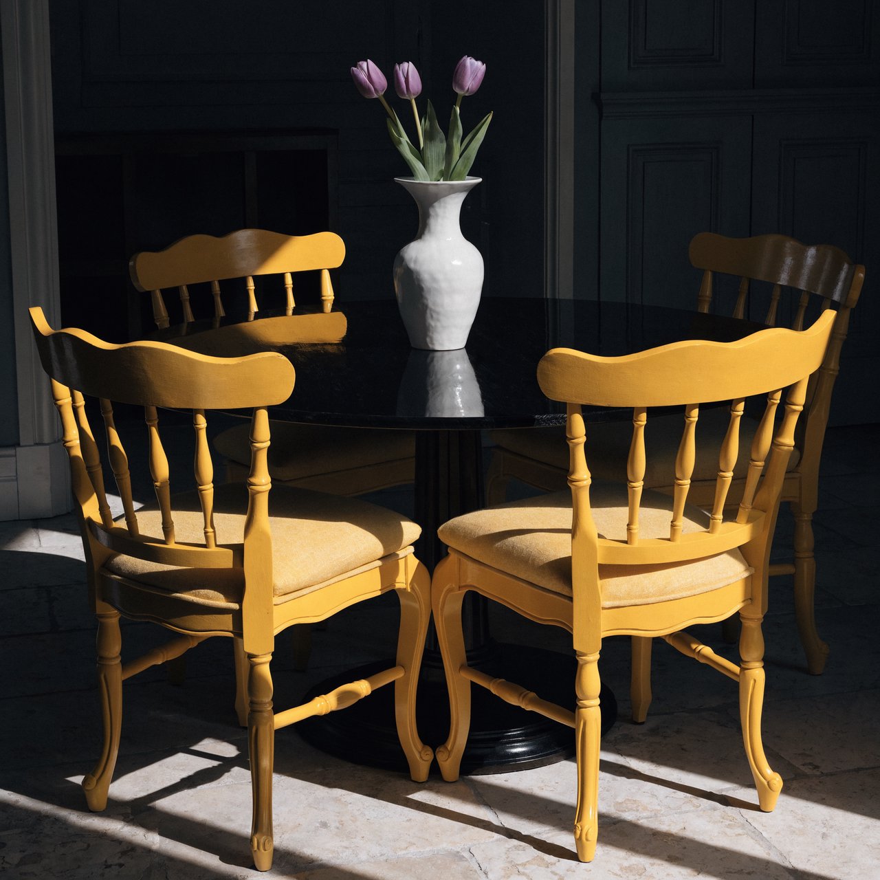 Exactly four yellow wooden chairs surround a round black table: two chairs at the front, one on the left, and one on the right. A white vase with three purple tulips sits at the table center, nothing else. — Recraft v4 Pro