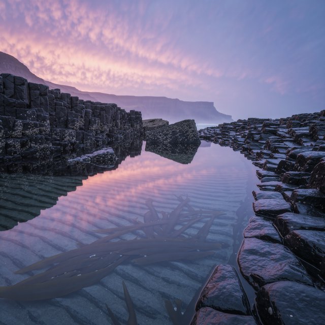At dawn on a rugged basalt shore, a tide pool mirrors clearing rain clouds; translucent water reveals rippled sand, barnacle-crusted rock, and blurred kelp below the surface, with slick foreground stone leading toward hazy coastal cliffs. — Kling v3 Omni