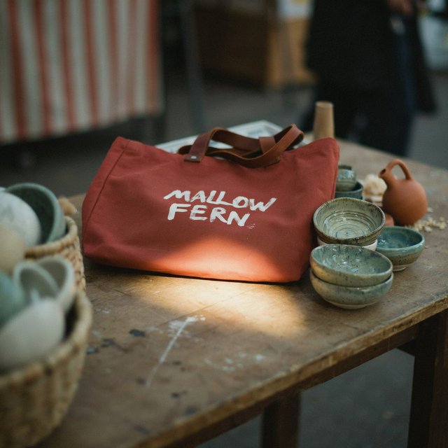 Sunlight pools over a pottery market table, where a rust-red canvas tote rests beside glazed bowls; "MALLOW FERN" is hand-painted in chunky white letters on its front, with no other legible writing. — Model 7