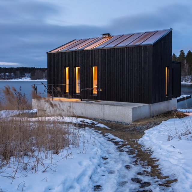 Exterior of a Scandinavian modernist lakeside sauna pavilion, blackened timber cladding, pale granite plinth, copper roof, winter blue-hour light glowing through narrow windows, viewed from a snowy reed-lined path, straight structural geometry and plausible construction. — ImagineArt 2