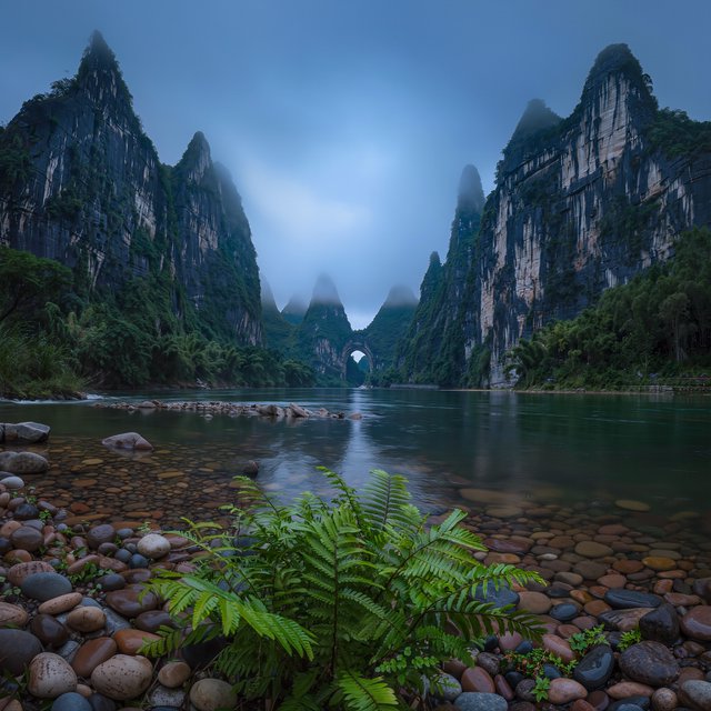 A limestone karst valley at blue hour after a light shower, towering pale cliffs rising above a clear river, low clouds snagging on pinnacles, slick ferns and rounded pebbles leading toward a distant stone arch. — Kling v3