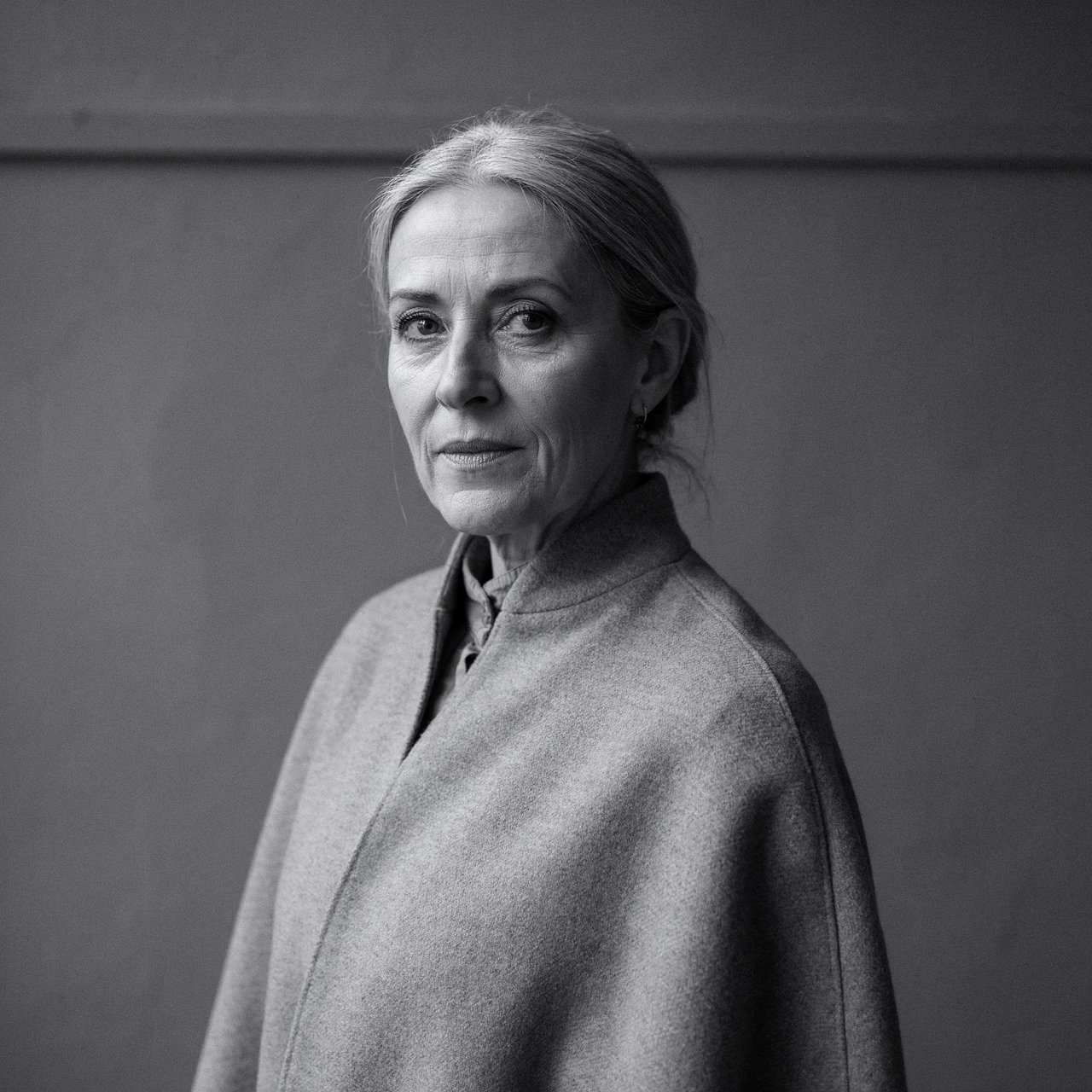 Under a narrow window beam, an adult woman in her late 50s wears a sculpted wool cape over a cotton blouse; her wry expression rests against a charcoal plaster backdrop in a black-and-white portrait, shaped by soft angled light. — Ideogram v4.5