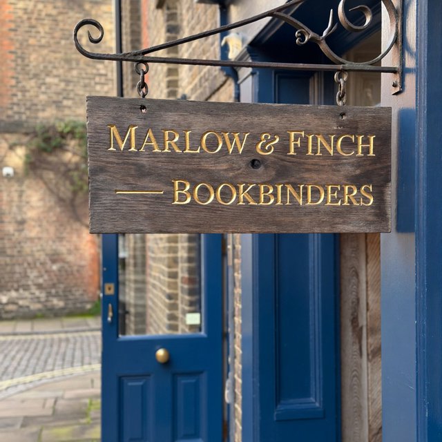 A hanging wooden shop sign that reads "MARLOW & FINCH — BOOKBINDERS" in gold serif letters, carved, above a blue door. — ImagineArt 2