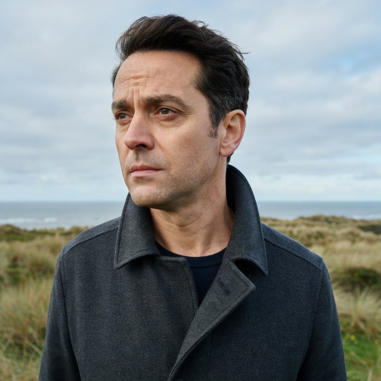 Wind lifts the hem of a charcoal wool coat as an adult man in his late 40s keeps a pensive gaze. The portrait frames him against a windswept coastal bluff beneath cool, cloud-filtered daylight. — Ideogram v4.5