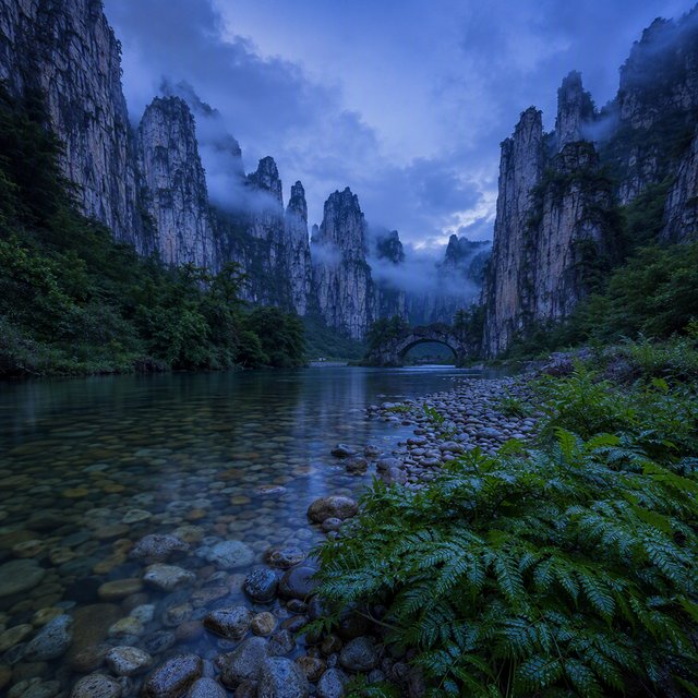 A limestone karst valley at blue hour after a light shower, towering pale cliffs rising above a clear river, low clouds snagging on pinnacles, slick ferns and rounded pebbles leading toward a distant stone arch. — GPT Image 2