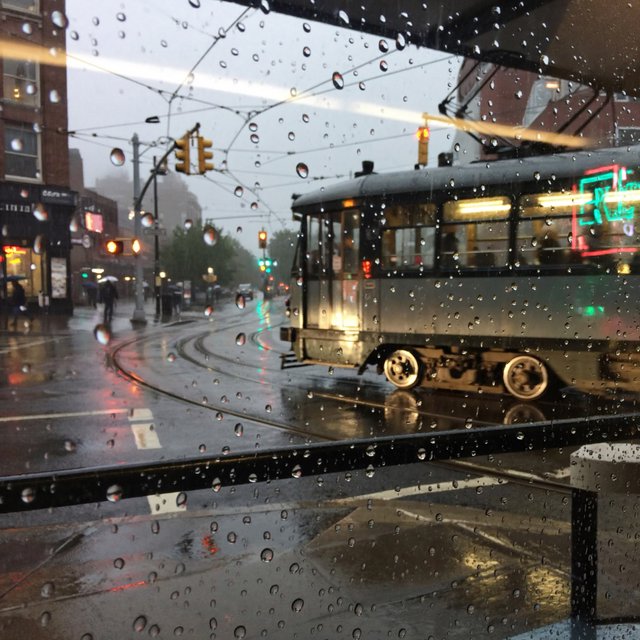 At a rain-darkened intersection, a tram grinds through a curve, its steel wheels shining beneath amber signal glow. A street-level news photo, shot on a 35mm lens with moderate depth of field, catches beads of water on the windows and slight wheel blur. — ImagineArt 2