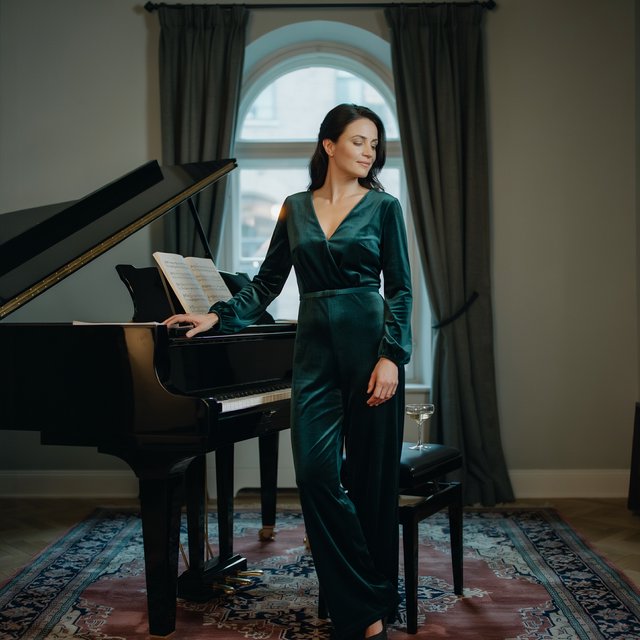 Editorial glamour photograph of an adult woman in a midnight-green velvet evening jumpsuit, standing naturally beside a grand piano in a softly lit salon, warm flattering light, relaxed hands, elegant posture, sophisticated fashion composition. — Ideogram v4