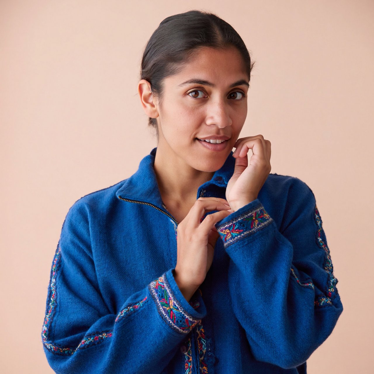 Fashion portrait of an adult Latina woman in her late 20s wearing a handwoven cobalt-blue cotton jacket, one hand touching its embroidered cuff, curious half-smile, against a pale peach studio backdrop, soft diffused window light shaping her face. — ImagineArt 2