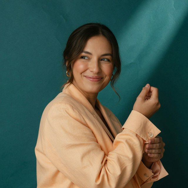 Portrait photograph of an adult woman in her early 30s wearing a pale apricot linen blazer, her hands gently smoothing its cuff. She looks quietly delighted against a deep teal paper backdrop, illuminated by soft, angled studio light. — Grok Imagine Image Quality