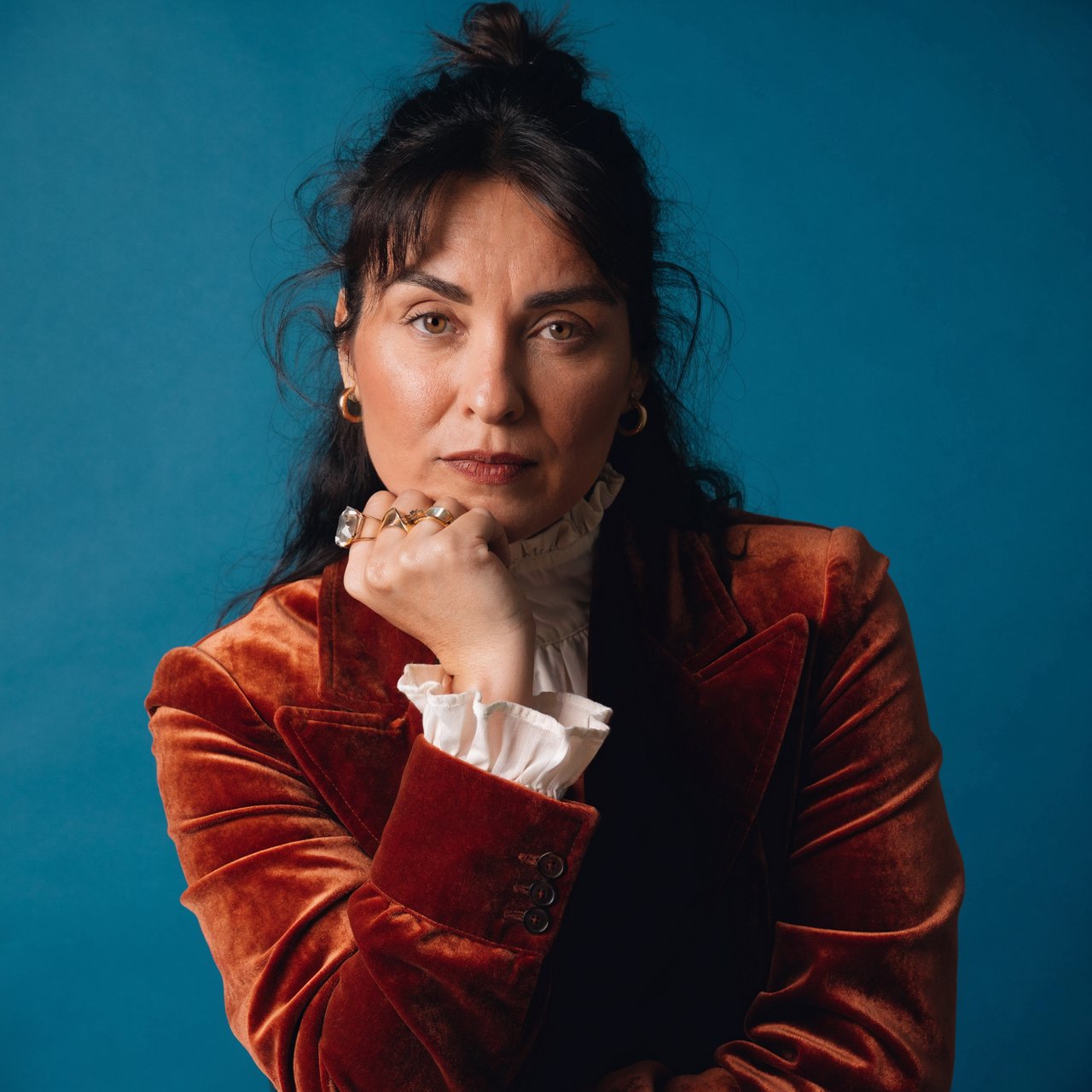 Portrait of an adult woman in her 30s wearing a rust-colored velvet jacket over an ivory blouse, one hand resting beneath her chin, thoughtful expression, deep teal studio backdrop, soft side lighting and gentle cheek highlights. — Recraft v4 Pro