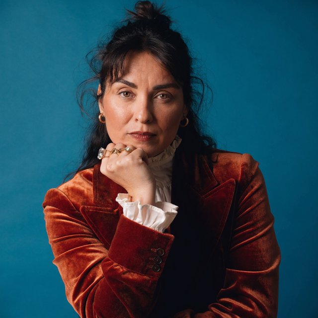 Portrait of an adult woman in her 30s wearing a rust-colored velvet jacket over an ivory blouse, one hand resting beneath her chin, thoughtful expression, deep teal studio backdrop, soft side lighting and gentle cheek highlights. — Recraft v4 Pro