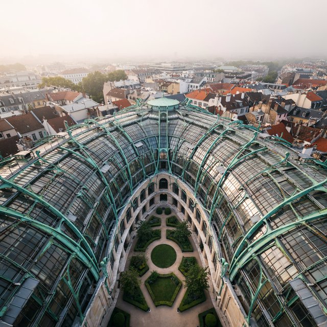 From a nearby rooftop, an Art Nouveau conservatory curves around a planted courtyard, its green copper ribs, clear glass, and pale stone base catching hazy morning light; orderly rooflines and believable supports show from above. — Ideogram v4.5