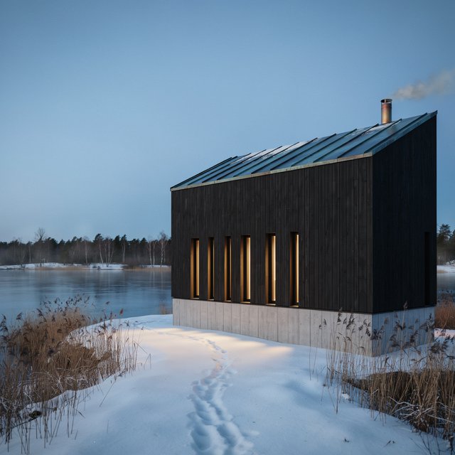 Exterior of a Scandinavian modernist lakeside sauna pavilion, blackened timber cladding, pale granite plinth, copper roof, winter blue-hour light glowing through narrow windows, viewed from a snowy reed-lined path, straight structural geometry and plausible construction. — Ideogram v4