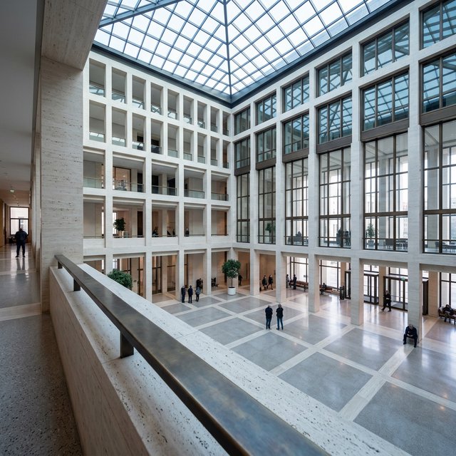 A soaring civic atrium in contemporary Italian Rationalist style pairs pale travertine, bronze mullions, and terrazzo beneath a glazed roof; cool morning light falls from above, viewed from a mezzanine, with aligned bays and credible supports. — Nano Banana 2.1