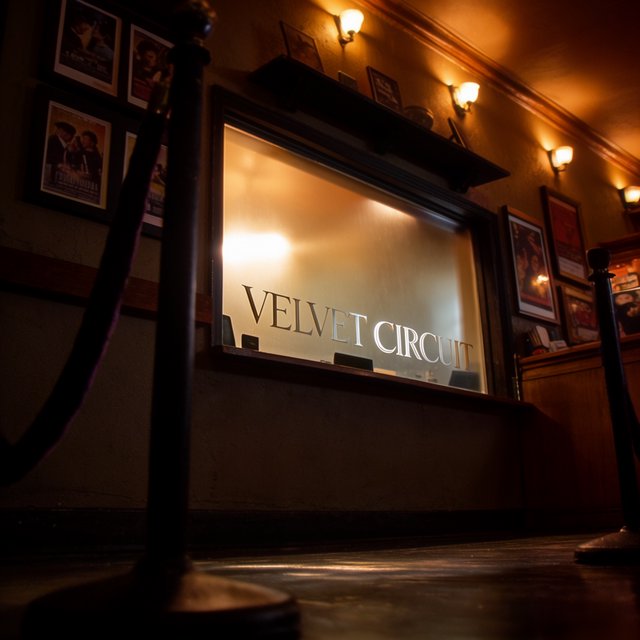 A narrow neighborhood cinema lobby glows with amber sconces, velvet ropes, and framed film stills; "VELVET CIRCUIT" is etched in elegant silver lettering across the frosted glass ticket-booth window. — Krea 2 Large