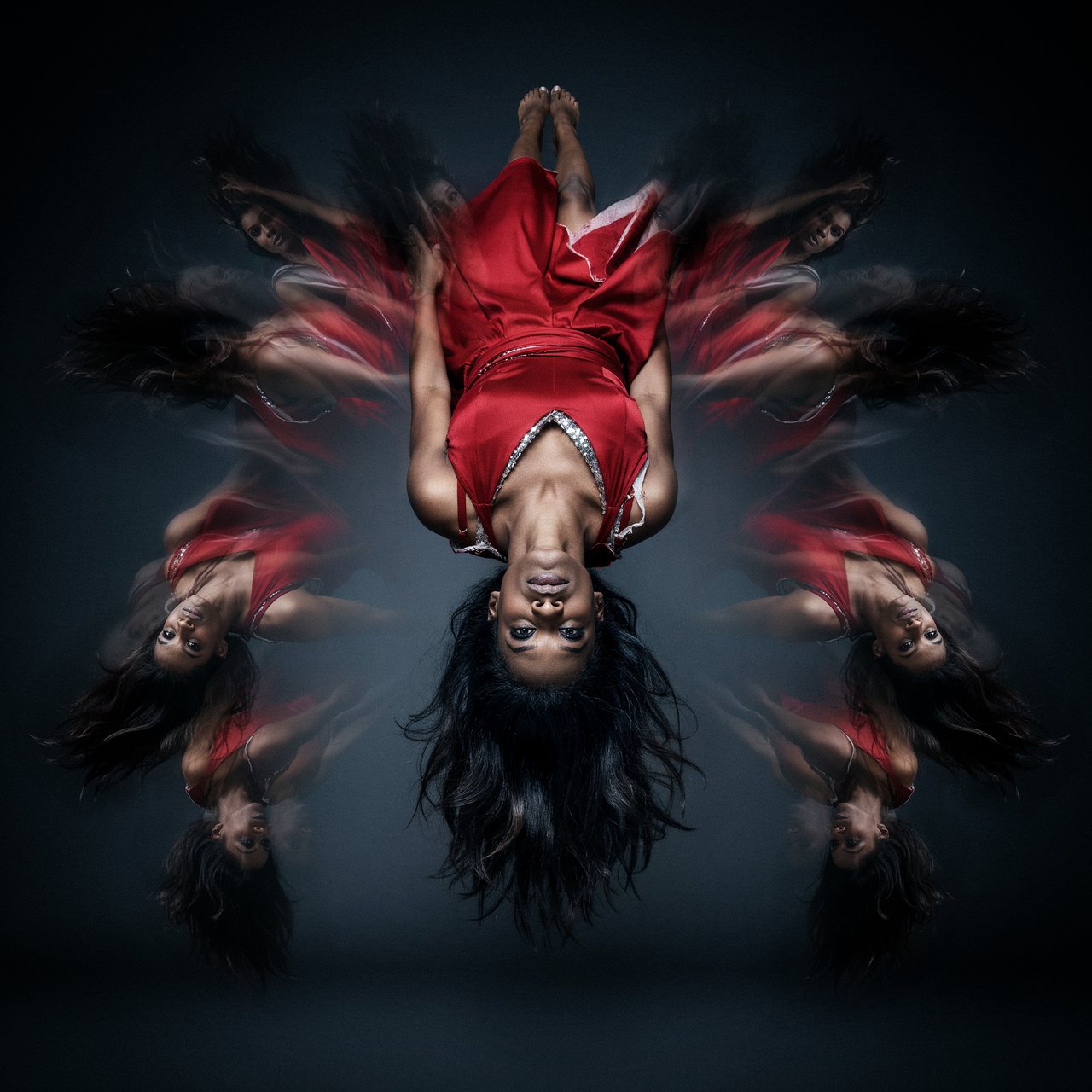 Image of a pop-dance African-American diva. The woman hangs completely inverted in mid-air, head pointing toward the floor and legs toward the ceiling. Her huge hair falls and spreads dramatically in the opposite direction, while her expression remains cool and composed. Her red gown is turned inside out, exposing its sparkling lining, seams, and construction. To push the surrealism further, a second, reversed version of her silhouette appears to peel outward from her original form, like a three-dimensional reflection escaping its owner. Multiple copies of the woman circle around a central point, each rotated to a different angle. Their flowing hair and red gowns form a pinwheel or circular vortex, creating the impression that one woman is spinning endlessly around herself. — Ideogram v4