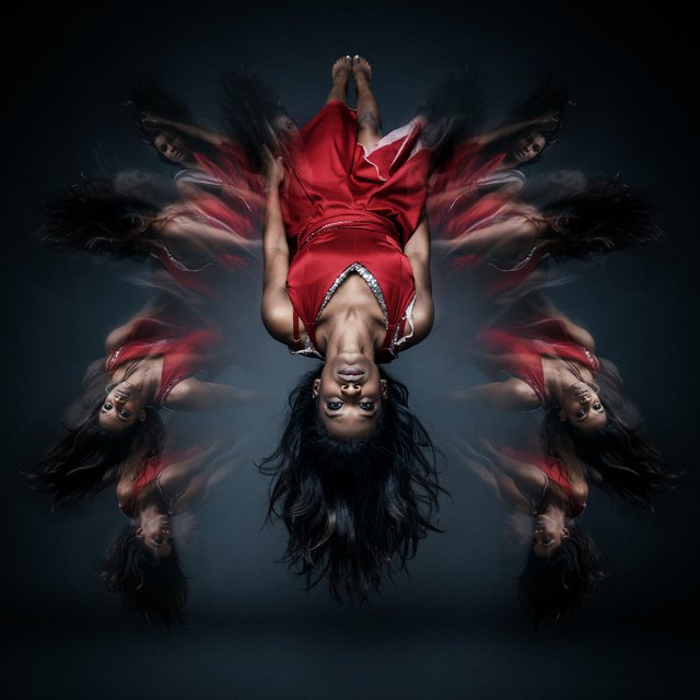 Image of a pop-dance African-American diva. The woman hangs completely inverted in mid-air, head pointing toward the floor and legs toward the ceiling. Her huge hair falls and spreads dramatically in the opposite direction, while her expression remains cool and composed. Her red gown is turned inside out, exposing its sparkling lining, seams, and construction. To push the surrealism further, a second, reversed version of her silhouette appears to peel outward from her original form, like a three-dimensional reflection escaping its owner. Multiple copies of the woman circle around a central point, each rotated to a different angle. Their flowing hair and red gowns form a pinwheel or circular vortex, creating the impression that one woman is spinning endlessly around herself. — Ideogram v4