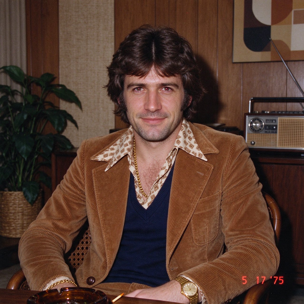 Create an image of a man in the quintessential style of 1975. Style his hair, clothing, accessories, and surroundings to reflect the distinctive look of the mid-1970s—featuring bold silhouettes, eye-catching accessories, layered details, and characteristic colors and textures. Give the image the feel of an authentic 1975 photograph, complete with analog film grain, faded colors, direct flash lighting, and a slight soft-focus effect. Add a period-accurate, red-orange date stamp in a 1970s style in one of the bottom corners. No recognizable posters. No modern objects or text. — Seedream 5.0 Flash