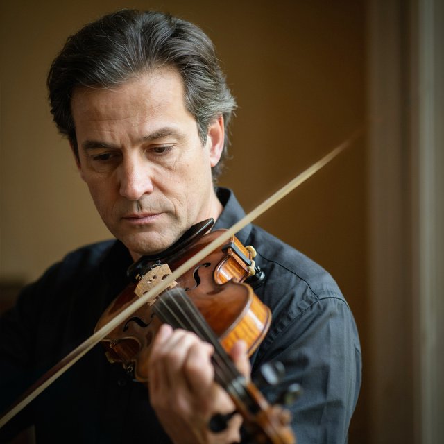 With a violin tucked close, an adult man in his early 50s draws a bow, his hands prominent, wearing a charcoal silk shirt; his intent expression is framed against a warm umber backdrop in soft amber window light, in a close portrait photograph. — Ideogram v4