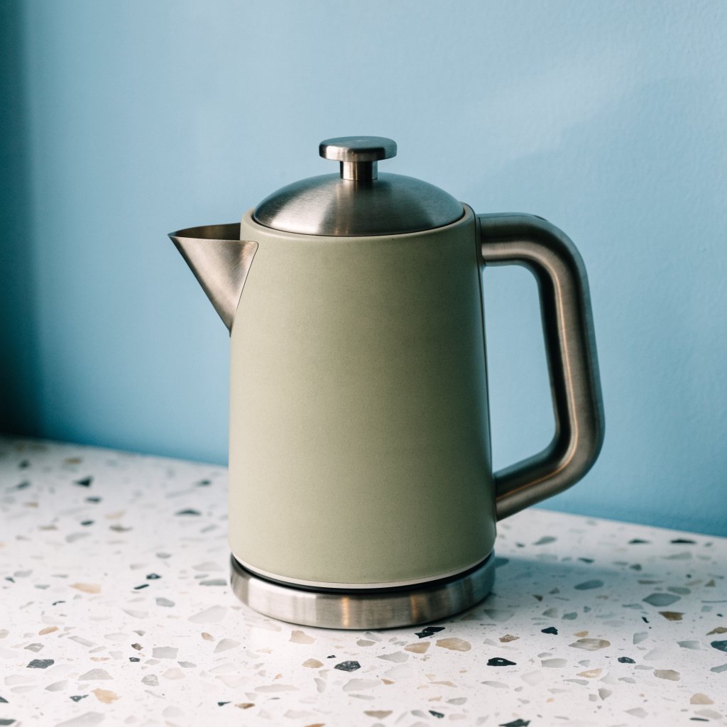 Commercial product photograph of an unbranded electric kettle made from matte sage ceramic and brushed stainless steel, resting on a white terrazzo surface against a pale blue background, with soft window light from the left and a delicate grounding shadow. — Recraft v4
