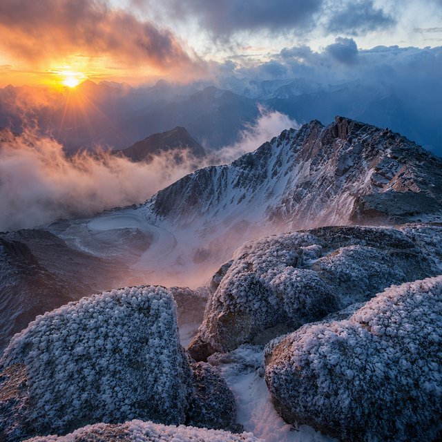 Wind combs a high granite ridge above a glacial cirque at sunrise, beneath clearing snow squalls; granular rime crusts foreground boulders while successive peaks dissolve into blue cloud shadow. — GPT Image 2