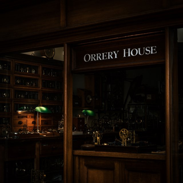 Inside a quiet clockmaker’s workshop, brass gears and tiny tools gleam beneath green-shaded lamps; "ORRERY HOUSE" is engraved in crisp serif letters on a dark walnut sign above the repair counter. — Krea 2 Large