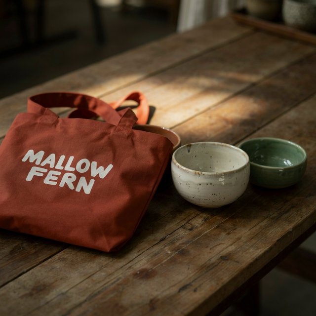 Sunlight pools over a pottery market table, where a rust-red canvas tote rests beside glazed bowls; "MALLOW FERN" is hand-painted in chunky white letters on its front, with no other legible writing. — Model 8