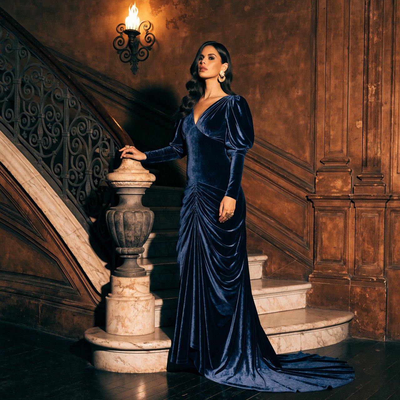 Editorial glamour photograph of an adult woman in a midnight-blue velvet evening gown, standing naturally beside a curved staircase in an old theatre, warm flattering sconce light, relaxed shoulders, poised expression, refined cinematic elegance. — Recraft v4 Pro