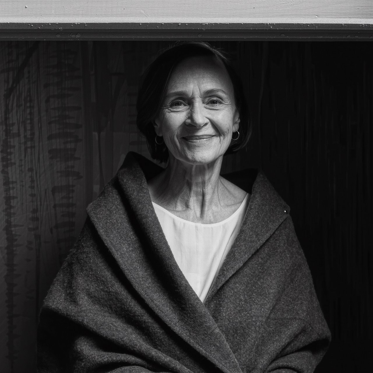 Under a narrow window beam, an adult woman in her late 50s wears a sculpted wool cape over a cotton blouse; her wry expression rests against a charcoal plaster backdrop in a black-and-white portrait, shaped by soft angled light. — Kling v3 Omni