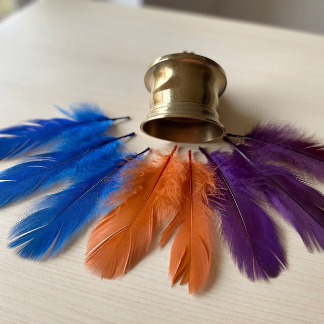 Exactly seven feathers lie on a cream tabletop: three blue on the left, two orange in the center, and two purple on the right. A brass bell sits above the orange pair, nothing else. — ImagineArt 2
