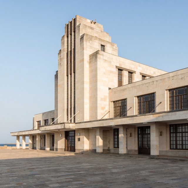 Exterior of a coastal Art Deco ferry terminal, cream limestone, dark bronze window frames, stepped geometric tower and sheltered arcade, late-morning sunlight casting sharp shadows, viewed from across a windswept stone forecourt, realistic architectural photography with precise lines and plausible structure. — ImagineArt 2