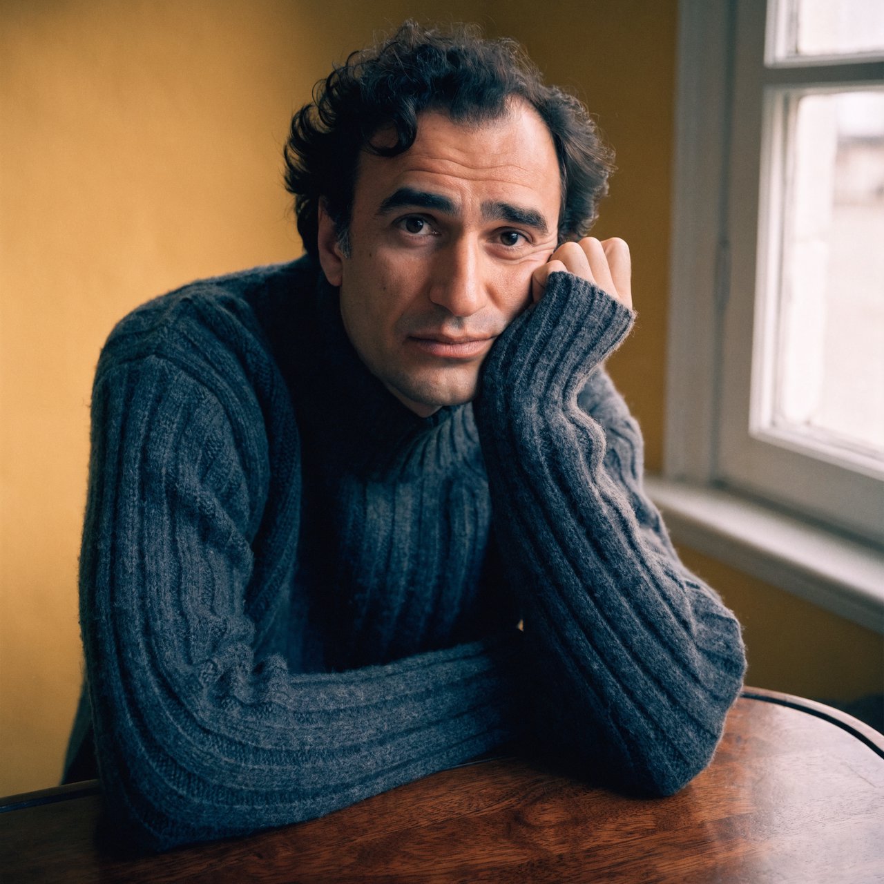 Editorial portrait of an adult man in his early 40s wearing a charcoal wool fisherman’s sweater, one hand loosely holding its ribbed cuff. His thoughtful, slightly tired expression rests against a muted ochre studio backdrop, shaped by gentle directional window light. — Recraft v4 Pro