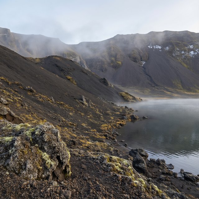 At first light, a rain-cleared volcanic caldera lake lies beneath thinning fog; pumice grains and damp lichen sharpen the near shore, while dark cinder slopes step back toward a distant rim. — Qwen Image 3.0