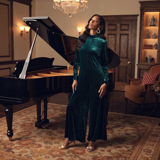 Editorial glamour photograph of an adult woman in a midnight-green velvet evening jumpsuit, standing naturally beside a grand piano in a softly lit salon, warm flattering light, relaxed hands, elegant posture, sophisticated fashion composition. — Recraft v4