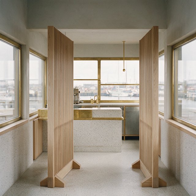 Inside an empty harbor café, restrained Danish modern design pairs ash slat screens, pale terrazzo, and brushed brass; overcast window light reaches the counter, viewed from the entrance, with aligned joinery and believable supports. — Model 20