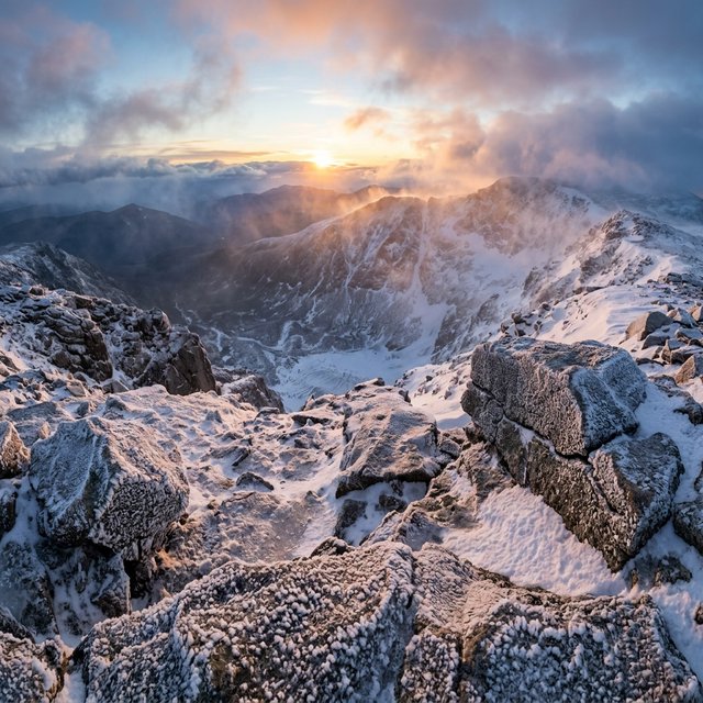 Wind combs a high granite ridge above a glacial cirque at sunrise, beneath clearing snow squalls; granular rime crusts foreground boulders while successive peaks dissolve into blue cloud shadow. — Nano Banana 2.1