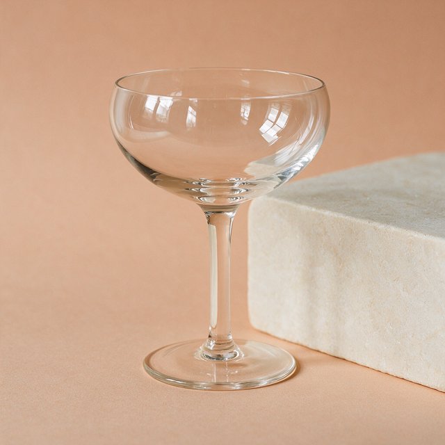 Beside a pale limestone block, an unbranded handblown cocktail coupe in clear soda-lime glass catches crisp window-shaped highlights; its curved bowl bends a muted apricot backdrop under broad diffused key light and narrow cool rim lighting, commercial product image, no lettering. — Seedream 5.0 Pro