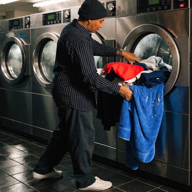 Candid documentary photograph of an adult laundromat attendant sorting warm clothes beside humming machines, cool fluorescent light, 50mm lens with moderate depth of field, loose lint clinging to a blue towel and faint condensation on the glass doors. — Recraft v4 Pro