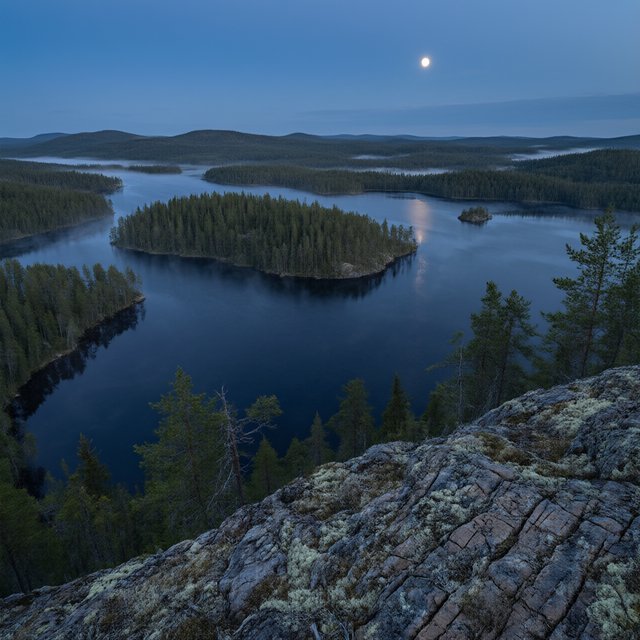 An expansive boreal lake beneath a clear, cold midnight sky, pale moonlight catching thin mist above dark water; spruce-covered islands recede toward distant ridges, while lichen and cracked granite sharpen the near shoreline. — Qwen Image 3.0