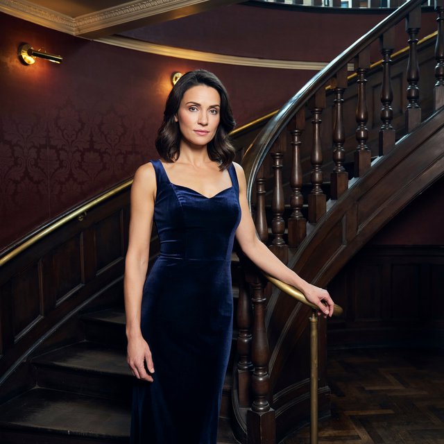 Editorial glamour photograph of an adult woman in a midnight-blue velvet evening gown, standing naturally beside a curved staircase in an old theatre, warm flattering sconce light, relaxed shoulders, poised expression, refined cinematic elegance. — Ideogram v4.5