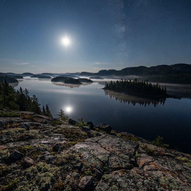 An expansive boreal lake beneath a clear, cold midnight sky, pale moonlight catching thin mist above dark water; spruce-covered islands recede toward distant ridges, while lichen and cracked granite sharpen the near shoreline. — Nano Banana 2 Lite
