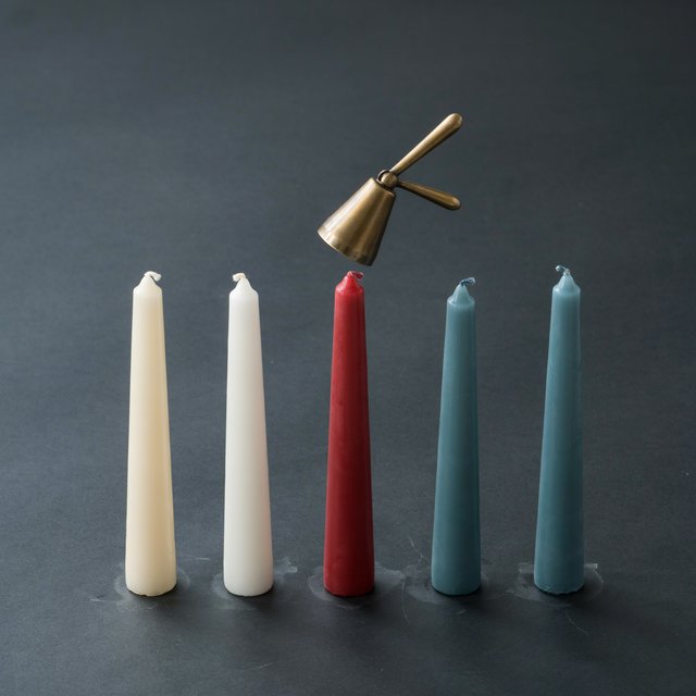 Exactly five candles stand on a dark slate tabletop: two ivory candles on the left, one red candle in the center, and two teal candles on the right. A brass snuffer sits above the red candle, nothing else. — Ideogram v4.5