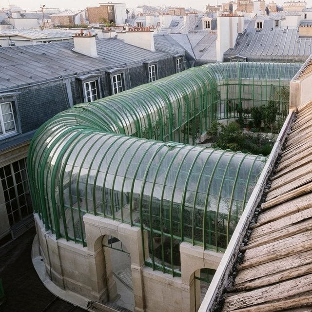 From a nearby rooftop, an Art Nouveau conservatory curves around a planted courtyard, its green copper ribs, clear glass, and pale stone base catching hazy morning light; orderly rooflines and believable supports show from above. — Krea 2 Large