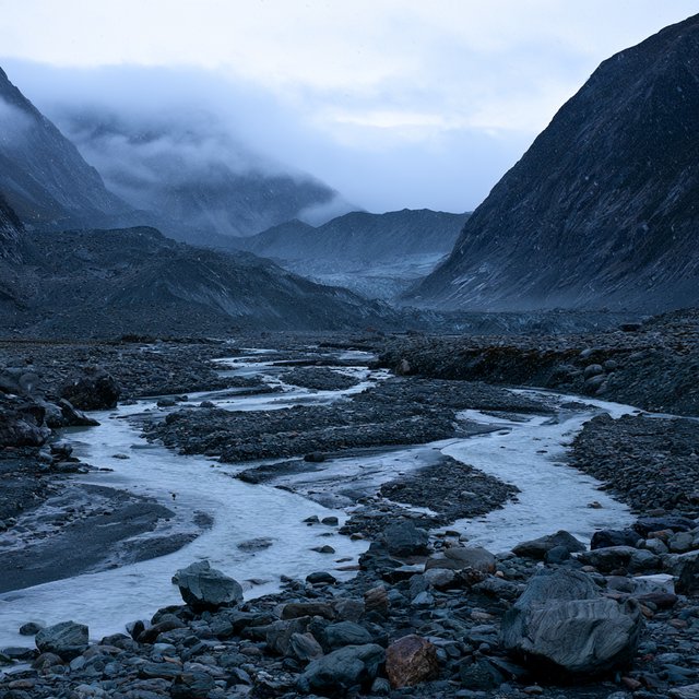 Beyond a braided glacier-fed stream, a steep moraine valley opens beneath clearing dawn snow flurries; gritty ice grains and water-polished stones sharpen the near bank, while layered ridges fade into cloud. — Model 13