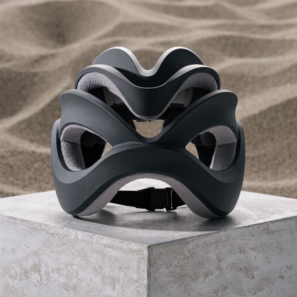 Commercial product photograph of an unbranded folding bicycle helmet made from matte charcoal polymer with soft gray padding, resting on a honed concrete plinth against a muted sand background, illuminated by a broad overhead softbox and gentle rim light. — Recraft v4