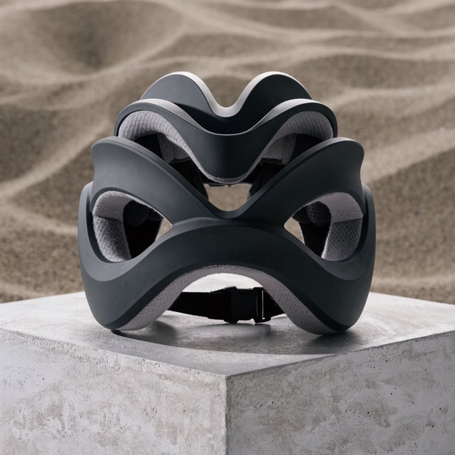 Commercial product photograph of an unbranded folding bicycle helmet made from matte charcoal polymer with soft gray padding, resting on a honed concrete plinth against a muted sand background, illuminated by a broad overhead softbox and gentle rim light. — Recraft v4